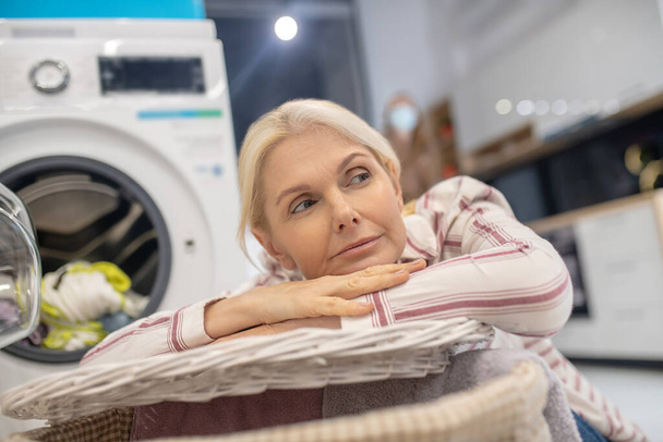 Blonde woman sitting near washing machine and looking thoughtful - 写真・画像