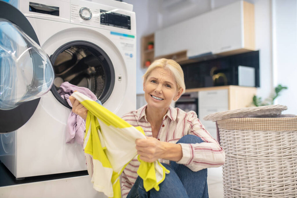 Femme au foyer blonde en chemise rayée et jeans assis près de la machine à laver - Photo, image