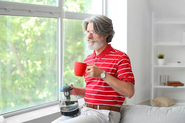Mature man with electric kettle and cup at home - Foto, Bild