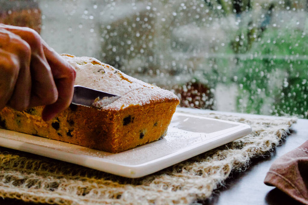 A hand cutting the freshly baked blueberry cake, rain behind the window creating a cozy autumnal atmosphere - Фото, изображение