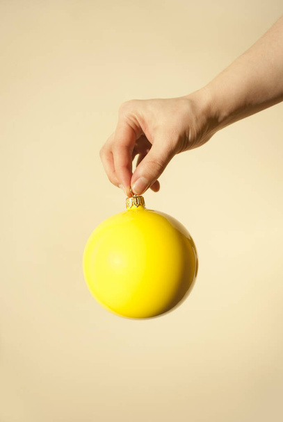 Ręcznie trzymając świąteczną żółtą bombkę dekoracji na beżowym tle. Minimalne pochodzenie noworoczne. Oświecające. Miejsce na tekst - Zdjęcie, obraz