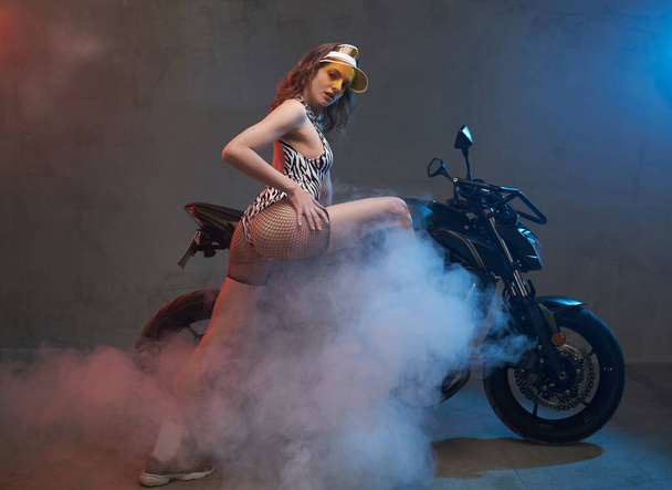 Beautiful female biker with cap poses with motorcycle in studio - Photo, Image