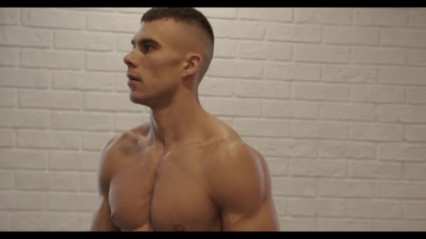 Mladý svalnatý muž s nahým trupem stojí na pozadí bílé cihlové zdi v tělocvičně a otáčí pohledy do kamery - Záběry, video