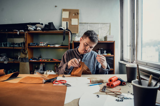 Sitting at a table in the workshop a young guy manually sews leather elements to each other - Photo, Image