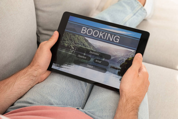 Man booking tickets online on sofa indoors, closeup. Travel agency concept - 写真・画像