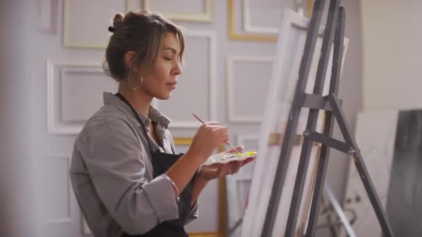 Plan moyen d'une femme métissée assise devant une toile blanche au chevalet dans un studio d'art moderne et lumineux faisant des coups avec un pinceau créant des œuvres d'art souriantes et appréciant le processus - Séquence, vidéo