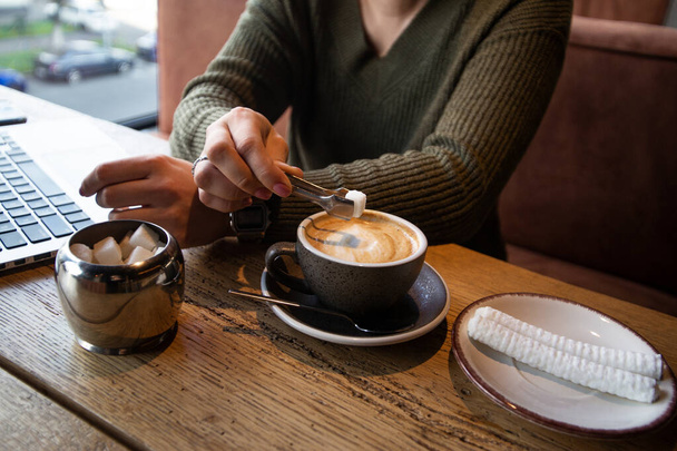 A zöld pulóveres nő hozzáad egy darab cukrot a kapucsínójához fogóval, miközben egy kávézóban dolgozik. Kényelmes kávézó koncepció. Vágott nézet laptop a háttérben. - Fotó, kép