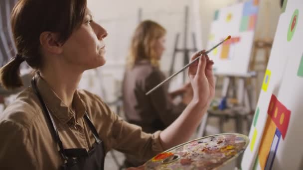 Medium close-up of young female arts student sitting in front of white canvas at easel holding palette and paintbrush creating abstract artwork at arts course - Footage, Video