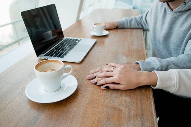 Womans hand with gold ring covers mans hand. Romantic date concept. Modern laptop and two empty coffee cups on wooden table. Casual style. Heterosexual couple good spending time. - Photo, Image
