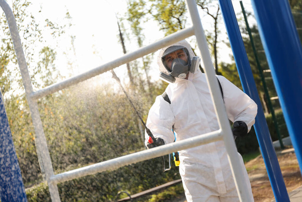 Man in hazmat suit spraying disinfectant on outdoor gym's equipment. Surface treatment during coronavirus pandemic - Photo, Image