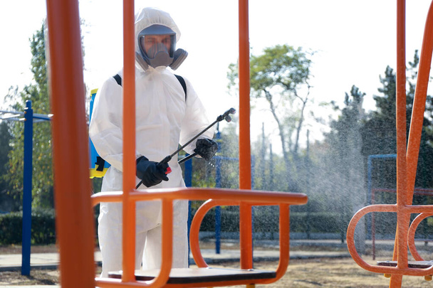 Man in hazmat suit spraying disinfectant onto swing at children's playground. Surface treatment during coronavirus pandemic - Photo, Image
