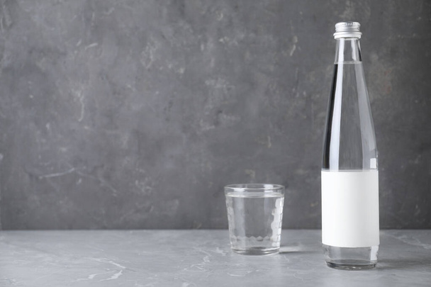 Bottle and glass with water on grey marble table, space for text - Фото, изображение