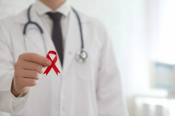 Doctor holding red ribbon on blurred background, closeup with space for text. AIDS awareness month - Foto, Imagem