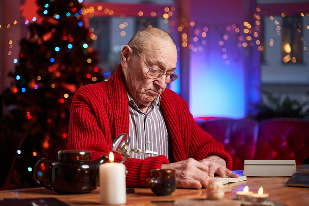Kopasz öregember szemüveggel ül az asztalnál és karácsonyi üdvözlőlapokat ír. - Fotó, kép