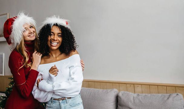 Τα Χριστούγεννα ενώνουν τη φυλετική ποικιλομορφία. Αφρικανικές και καυκάσιες νεαρές γυναίκες με καπέλο Αϊ Βασίλη στο σπίτι αγκαλιάζουν και κοιτάζουν την κάμερα. Μαύρη ζωή ύλη και την έννοια της ανοχής - Φωτογραφία, εικόνα