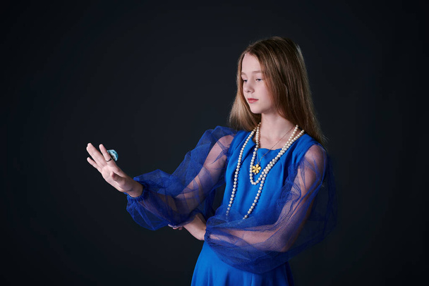 girl teenager in a blue dress tries on a ring on finger. studio. dark background - Photo, Image