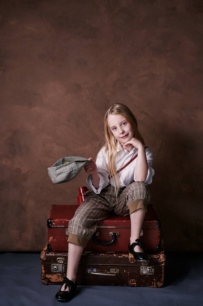 little girl sits on suitcases. vintage style photography. child in a white shirt, pants and a hat with a visor. studio. brown background - Foto, afbeelding
