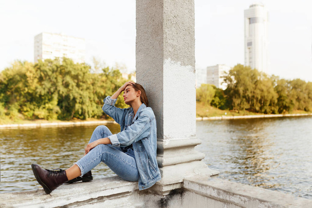 Ciddi bir bekar genç kızın profili, kot giymiş, nehrin kenarında oturmuş, başka yöne bakıyor.. - Fotoğraf, Görsel