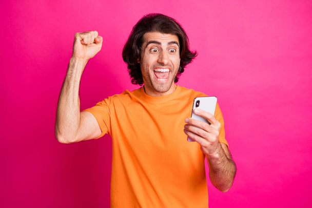 Photo of young man open mouth hold cellphone look screen fist up wear orange t-shirt isolated pink color background - Valokuva, kuva