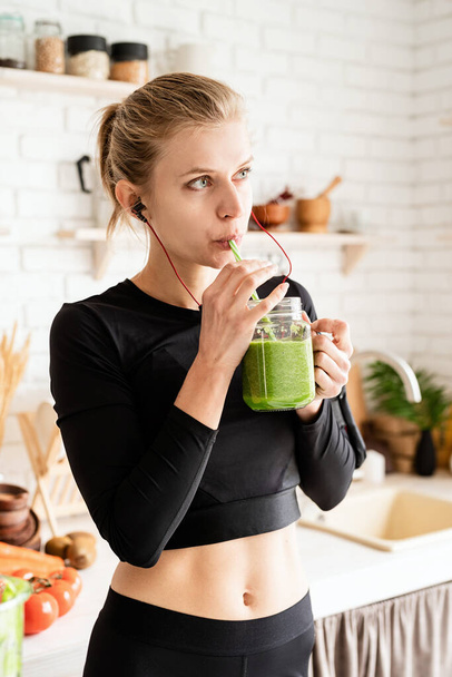 Sağlıklı beslenme, diyet konsepti. Genç sarışın kadın ev mutfağındaki kavanozdan yeşil smoothie içiyor. - Fotoğraf, Görsel