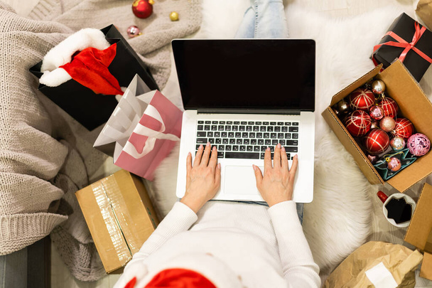 Připravuje se na vánoční večírek. Žena objednávající dárky a dekorace na notebooku, posezení mezi dárkovými krabicemi a balíčky, kopírovací prostor - Fotografie, Obrázek