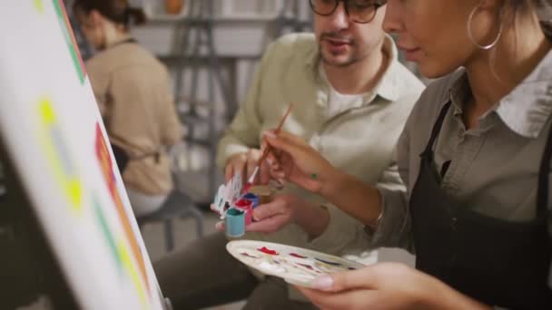 Zeitlupenaufnahme einer jungen Frau mit gemischter Rasse im Kunstunterricht, die Grundlagen abstrakter Malerei lernt, während sie dem männlichen Lehrer beim Laden von Pinsel und Gouachefarbe im hellen Atelier zuhört - Filmmaterial, Video