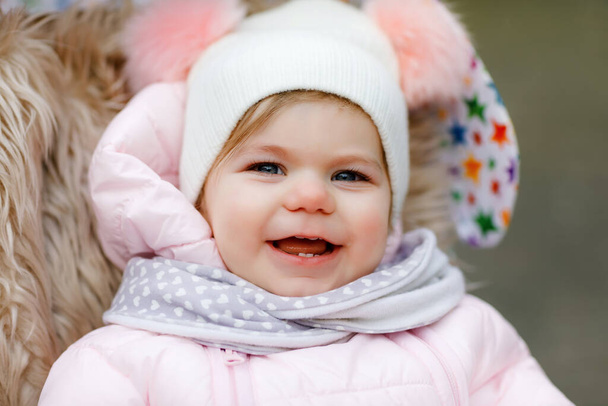 Söpö pieni kaunis tyttö istuu lastenvaunuissa tai rattaissa kylmänä syksyllä, talvella tai keväällä. Onnellinen hymyilevä lapsi lämpimissä vaatteissa, muodikas vauvan takki ja hattu. Sataa lunta - Valokuva, kuva