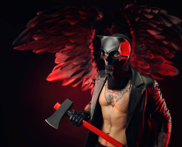 un hombre con una máscara de cráneo de demonio en una capa de cuero con un hacha sobre un fondo de alas negras y rojas - Foto, Imagen