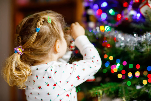 Adorable toddler girl in pajamas decorating Christmas tree with toy in cute hands. Little child in nightwear standing by Xmas tree. celebration of traditional family winter holiday - Foto, Imagen