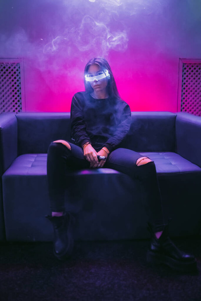 A young woman in glowing glasses sits under a neon light. Neon noir. - Foto, afbeelding