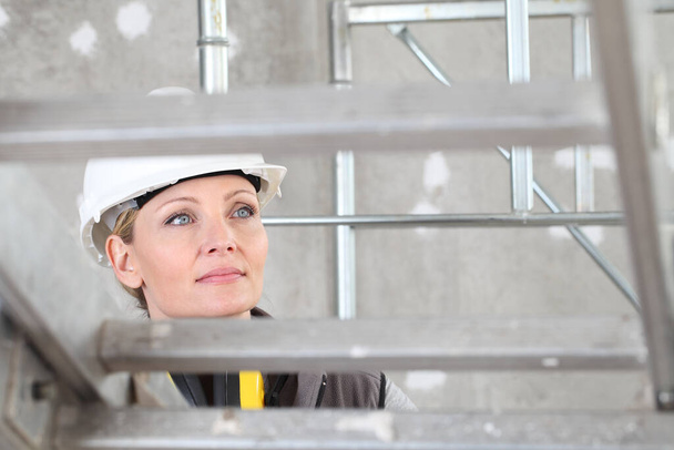 glimlachende vrouw bouwvakker op ladder met witte helm en gehoorbescherming koptelefoon op interieur bouwplaats achtergrond - Foto, afbeelding