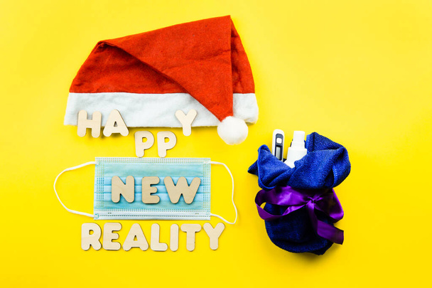 Santa hat, protective face mask and wooden letters HAPPY NEW REALITY on yellow background. - Photo, image