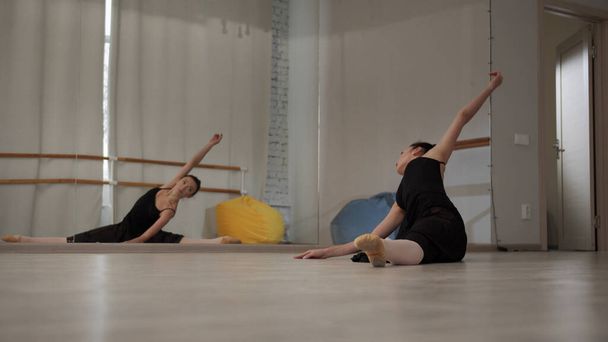 Voor een repetitie in een danszaal strekt een prachtige ballerina haar armen en benen uit terwijl ze op de vloer zit op een spleet voor een spiegel - Foto, afbeelding