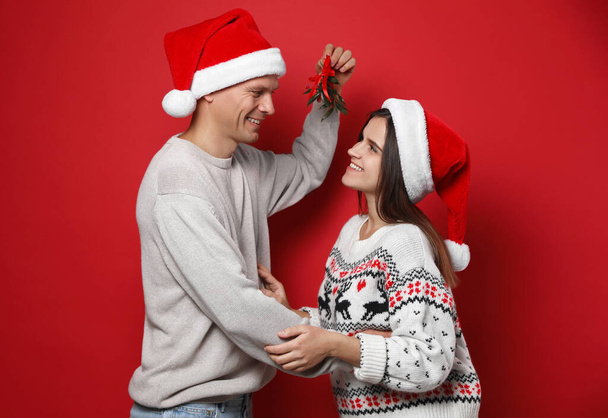 Happy couple in Santa hats standing under mistletoe bunch on red background - Foto, Imagen