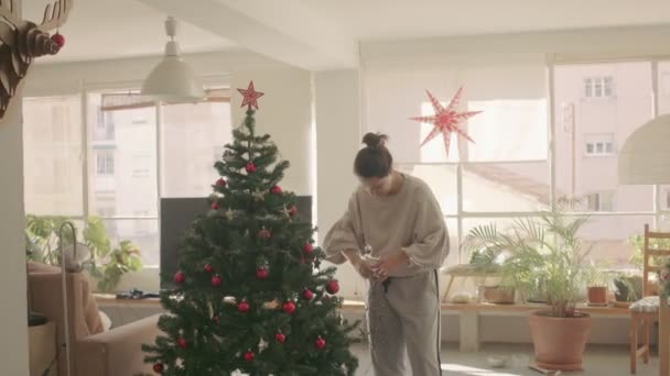 Donker haar Kaukasisch meisje versieren een kunstmatige kerstboom thuis met een slinger. Medium Wide Camera shot 4k videobeelden van hoge kwaliteit. - Video