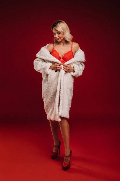 portrait of a smiling girl in a white fur coat and underwear on a red background. - Photo, Image