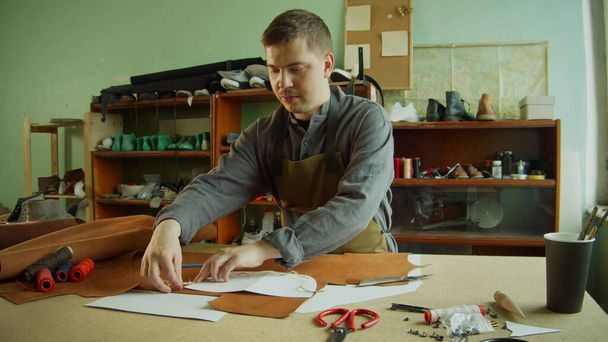 経験豊富な男性タナーは、ワークショップで作業台の上に茶色の革の大きな部分にバッグ要素のレイアウトを作ります. - 写真・画像