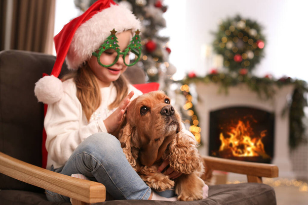 Cute little girl in Santa hat and Christmas glasses with English Cocker Spaniel at home - Photo, Image