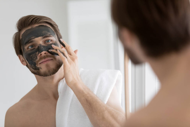 Peaceful young man applying black charcoal clay mask on face. - Φωτογραφία, εικόνα