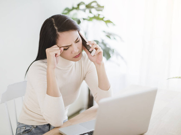 напряженная молодая женщина работает на ноутбуке и разговаривает по телефону в домашнем офисе - Фото, изображение