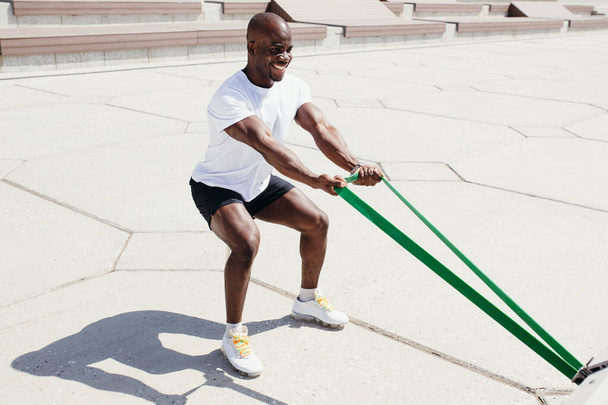 Afro American man in white t-shirt and black shorts doing exercise on his hands squatting with elastic fitness tape outside. - Foto, Imagem