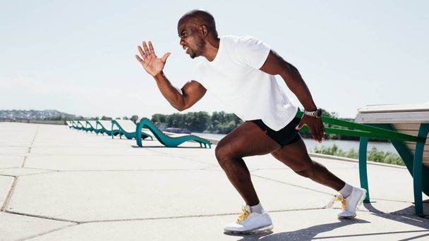 Black guy in white t-shirt and black shorts exercising and screaming outside with elastic tape. - Foto, Imagen