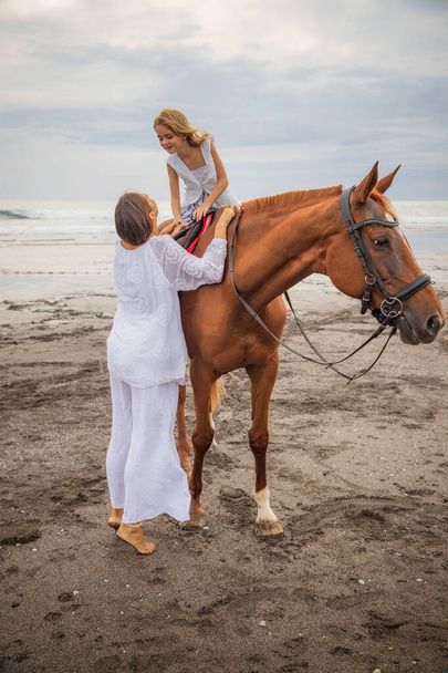 ビーチで乗馬。かわいい茶色の馬の上の女の子。彼女の母親は近くに立っている。動物への愛。母と娘は一緒に時間を過ごす。野外活動。母の日だ。インドネシアのバリ - 写真・画像