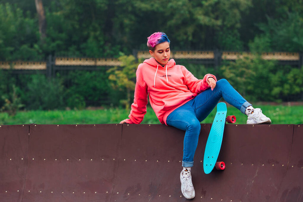 Portrait of a trendy pretty young girl with short colored hair and nose piercing dressed in pink hoodie and jeans sitting next to the skateboard court with her blue plastic skateboard. Copy space - Foto, afbeelding