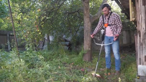 A kertész a vidéki ház udvarán dolgozik fűnyíróval. Férfi farmer vadfüvet nyír elektromos kaszával a farmon. Fiatal férfi munkás füvet nyír, nyírót használ. Benzinkefe-vágó - Felvétel, videó