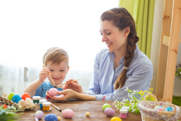Mother and her son painting colorful easter eggs. Family celebrating Easter. Parent and kid play indoors. Decorated home and spring flowers. - Фото, зображення