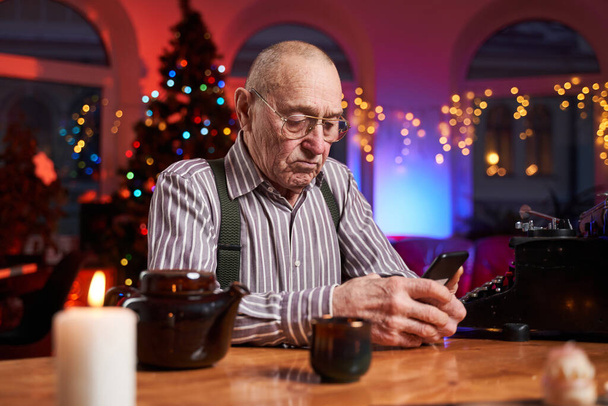 Radosny starszy człowiek za pomocą telefonu komórkowego siedzi przy stole w pokoju - Zdjęcie, obraz