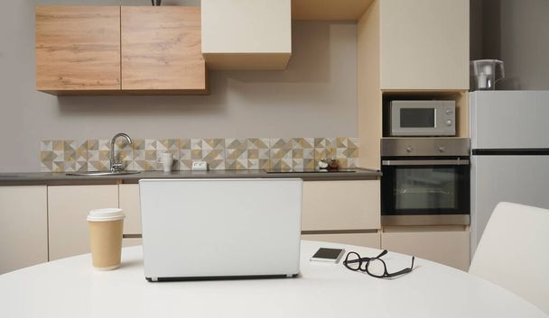 silver Laptop, mobile phone , glasses and plastic coffee cup on white wooden  table with kitchen background. no people. - Foto, afbeelding