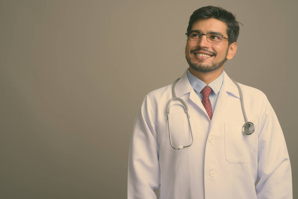 Στιγμιότυπο του νεαρού όμορφου γενειοφόρου Πέρση άνδρα γιατρό φορώντας γυαλιά με γκρι φόντο - Φωτογραφία, εικόνα