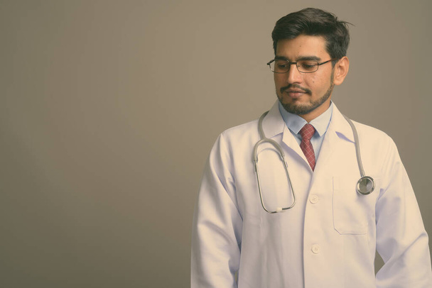 Στιγμιότυπο του νεαρού όμορφου γενειοφόρου Πέρση άνδρα γιατρό φορώντας γυαλιά με γκρι φόντο - Φωτογραφία, εικόνα